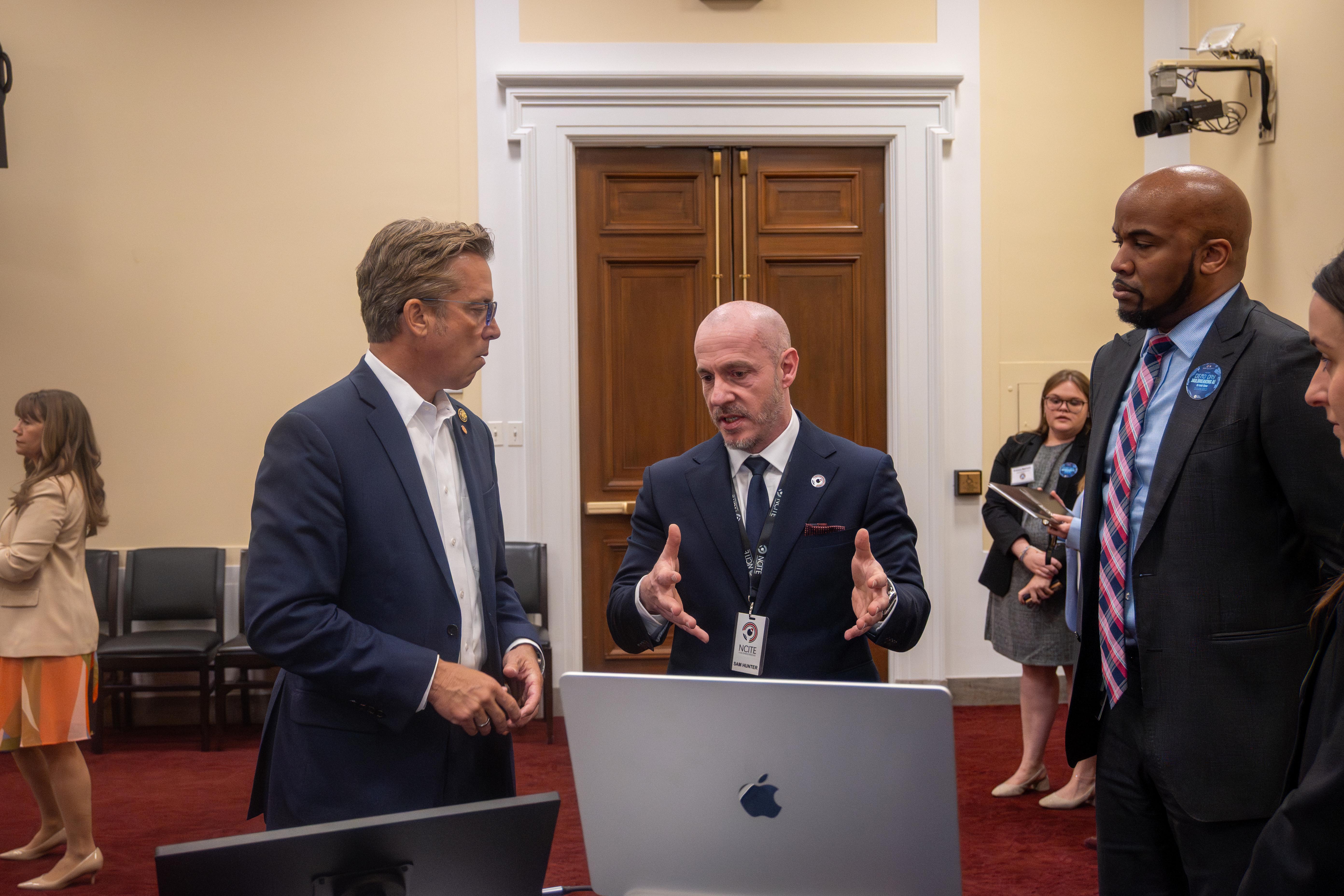 Three men in suits stand around a laptop as one gestures while explaining something during a discussion in a formal room.