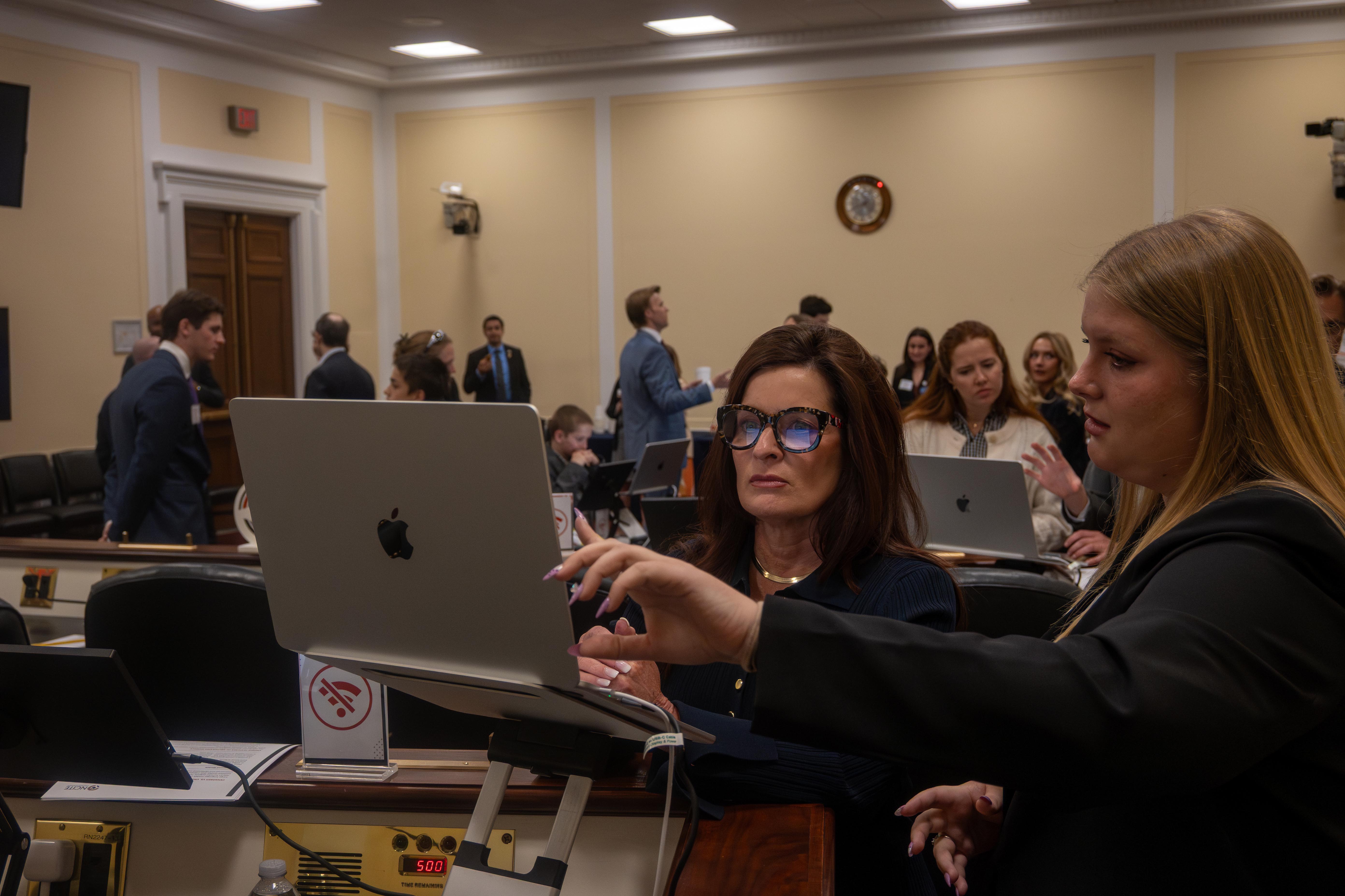 Two women in business attire collaborate at a laptop in a meeting room, with other attendees working and talking behind them.
