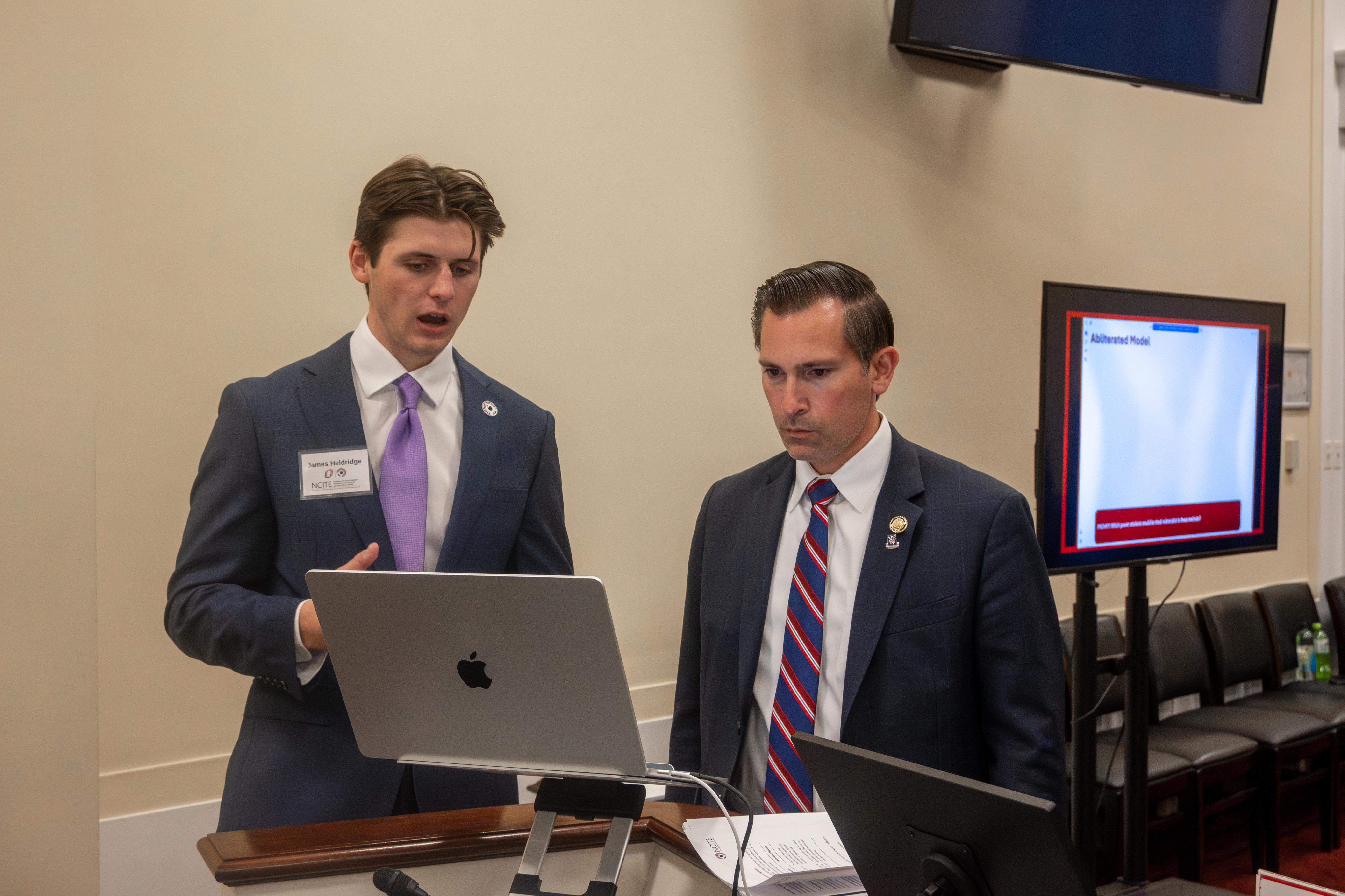 Two men in suits stand at a podium, focused on a laptop screen during a presentation in a conference room.