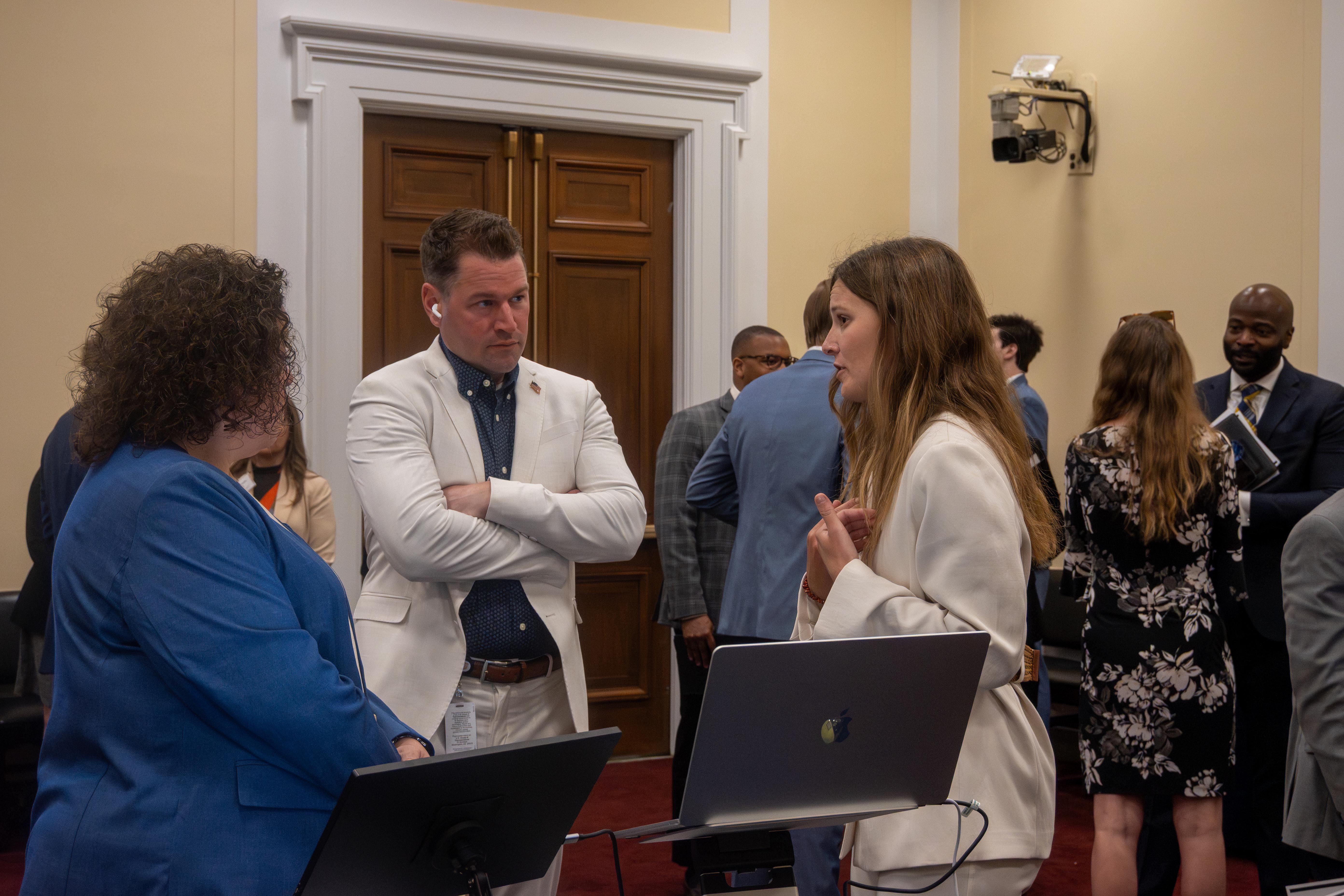 Three professionals in business attire stand in a meeting room, talking near a laptop on a stand while others converse in the background.