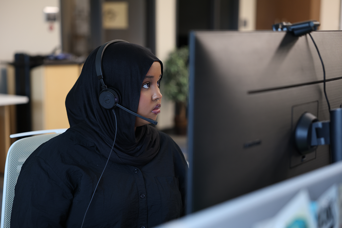 Hafso Ibrahim, wearing a black hijab and headset sits at a desk, focused on a computer monitor in an office setting. A webcam is mounted on top of the screen, and the background is softly blurred.