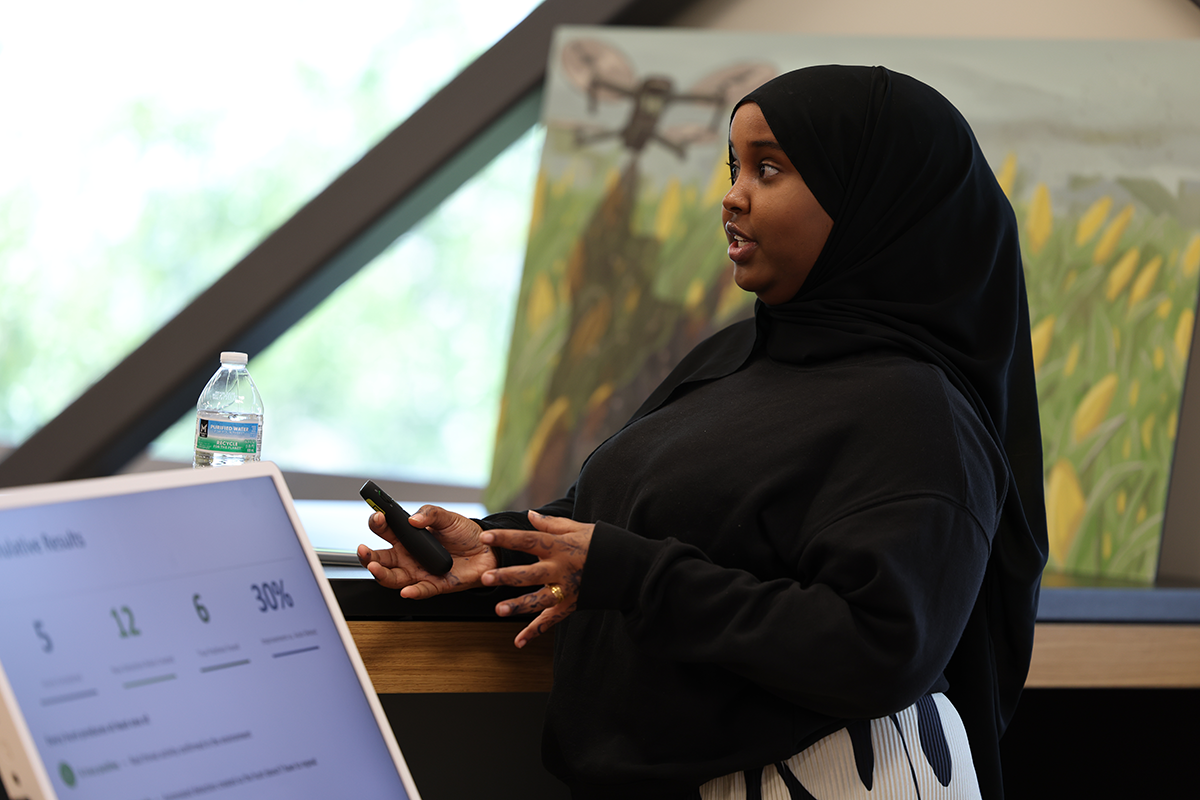Hafso Ibrahim, wearing a black hijab, gives a presentation while holding a remote control and gesturing with her hands. A presentation screen and water bottle are visible in the foreground, with artwork and large windows in the background.