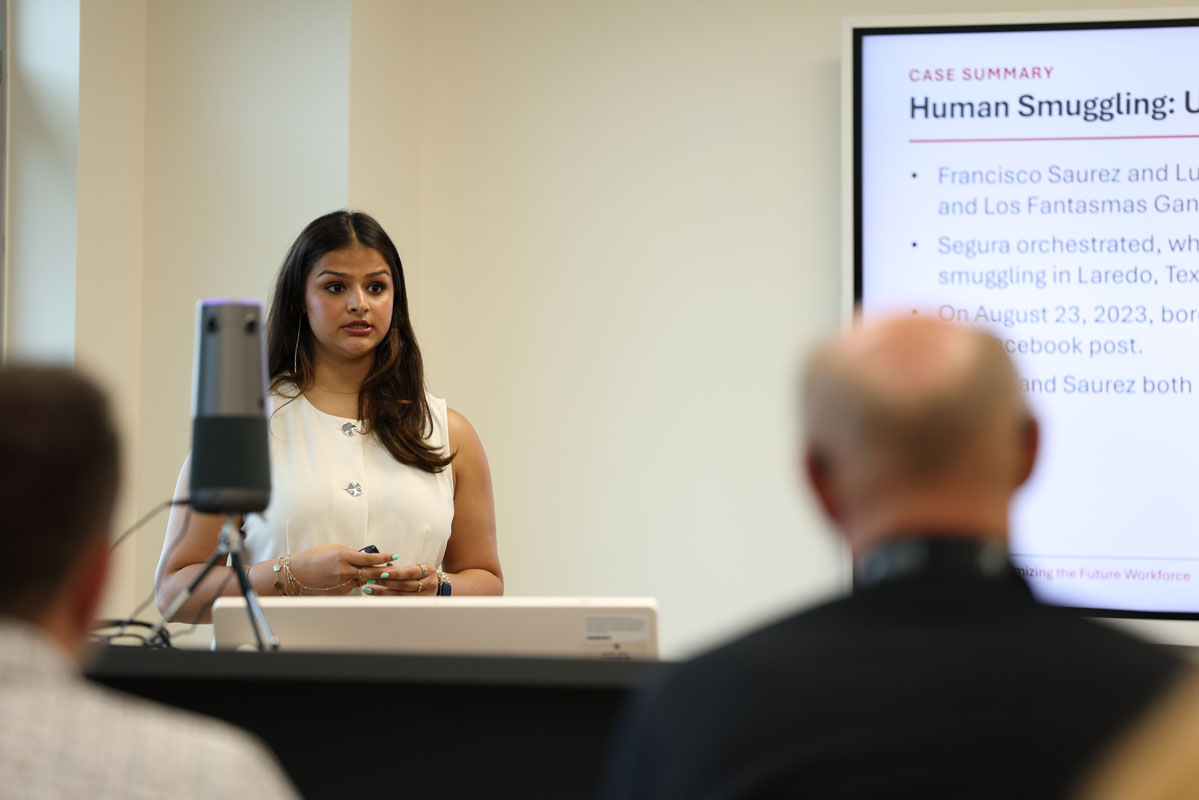 A presenter speaks at the front of a conference room while standing behind a podium. A presentation slide about a human smuggling case study is displayed on a large screen beside the speaker as audience members watch.
