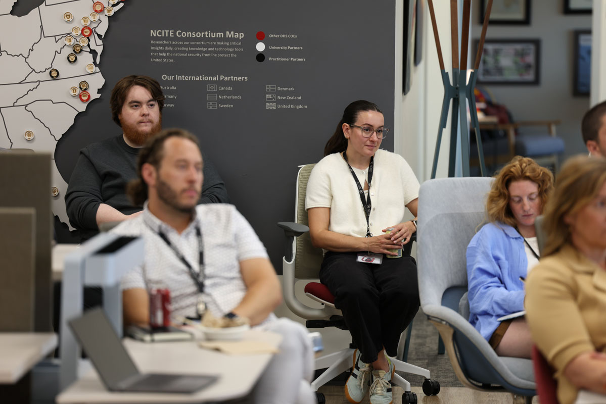 Attendees sit and listen during a presentation in an office meeting space. An NCITE consortium map is visible on the wall behind the audience, and several participants take notes while others watch the speaker.