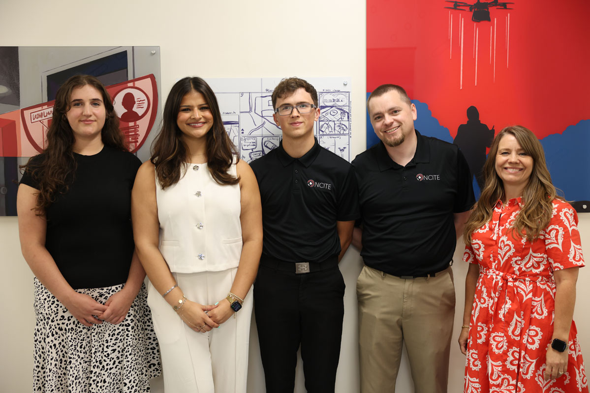 Five people stand together in an office in front of NCITE-themed artwork, posing for a group photo. Two people wear black NCITE polo shirts, while the others wear business or business-casual attire.