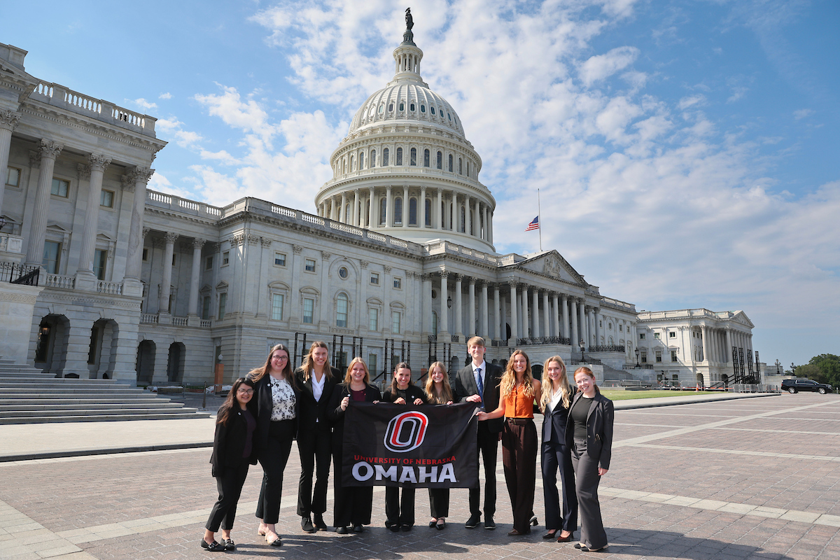 Ten NCITE students pose together in front of the U.S. Capitol in Washington, D.C., during a visit for congressional artificial intelligence demonstrations. The group stands smiling with the Capitol dome rising behind them under a blue sky with scattered clouds.