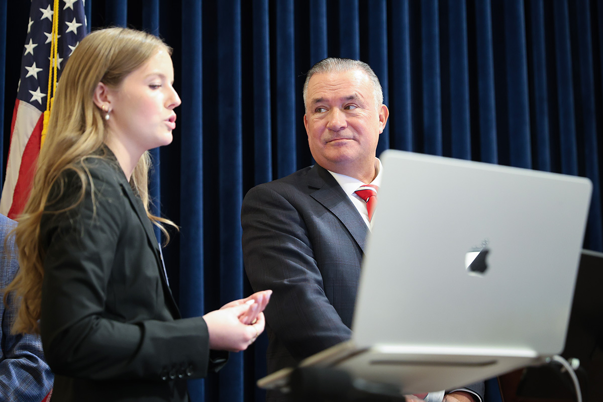 U.S. Rep. Don Bacon listens as an NCITE student researcher explains an artificial intelligence demonstration during a congressional event on Capitol Hill. Standing beside an open laptop, the student presents the research while Bacon looks on attentively, with a U.S. flag and the hearing room's blue curtains in the background.