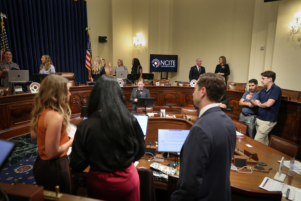 NCITE researchers, students, and congressional staff prepare for artificial intelligence demonstrations inside a U.S. House committee hearing room on Capitol Hill. Laptops are set up around the room as participants talk before the event, with an NCITE logo displayed on a monitor behind the committee dais.