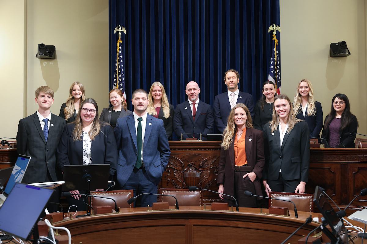 A group of 12 researchers, staff, and students stand with two congressional representatives in a U.S. House committee hearing room on Capitol Hill. The group poses in front of the committee dais beneath U.S. flags after an AI demonstration and briefing, with microphones, desks, and computer monitors visible in the foreground.