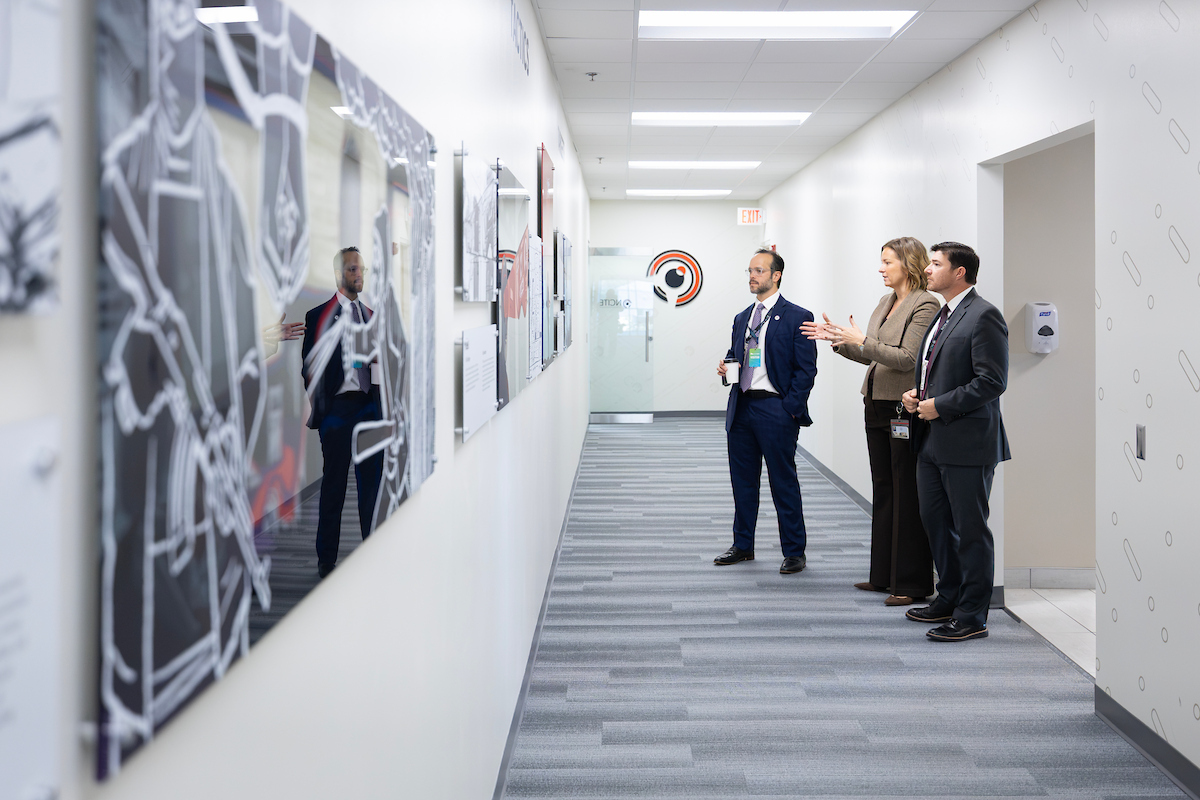 Three people gesture toward a wall display, tour a hallway featuring research exhibits at NCITE headquarters.