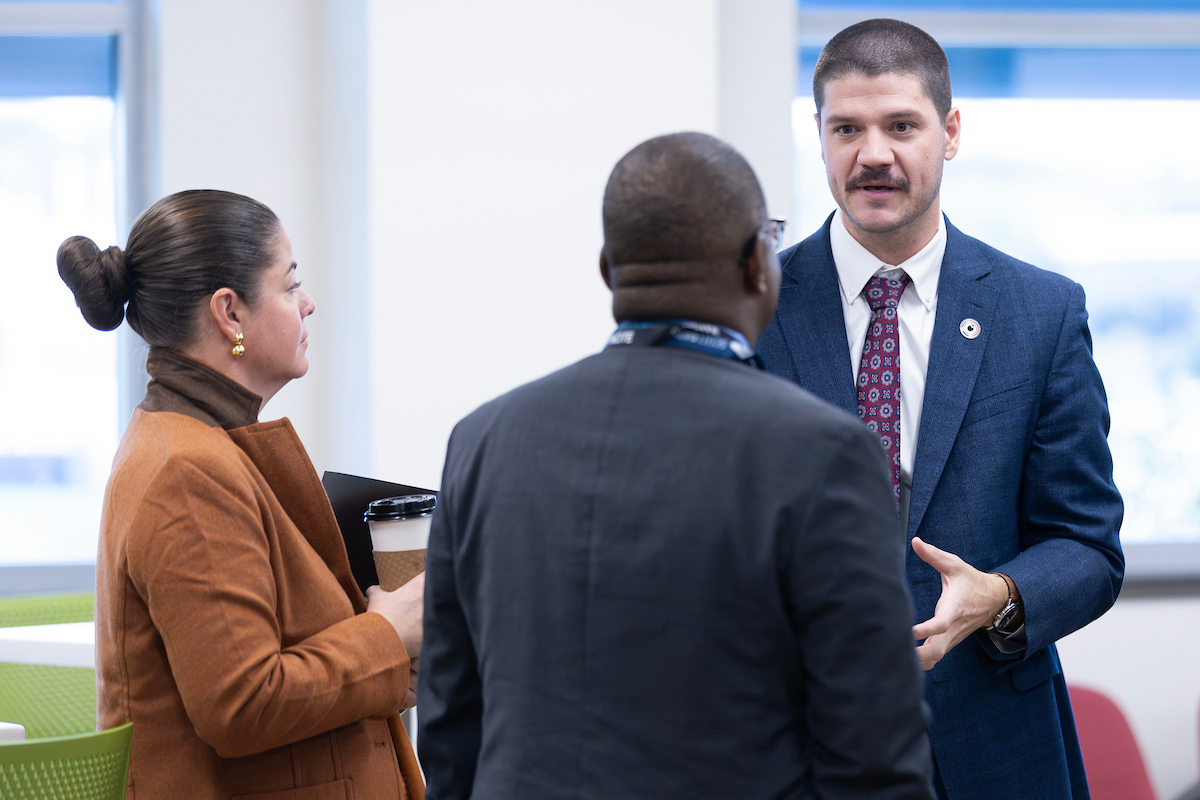 A man in a blue suit gestures while speaking with two NCITE visitors, including a woman holding a coffee cup, during a discussion at NCITE headquarters in Omaha.