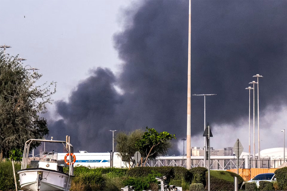 Thick plumes of black smoke rise above a fenced industrial facility in the United Arab Emirates, with trees, streetlights, and a small boat visible in the foreground.