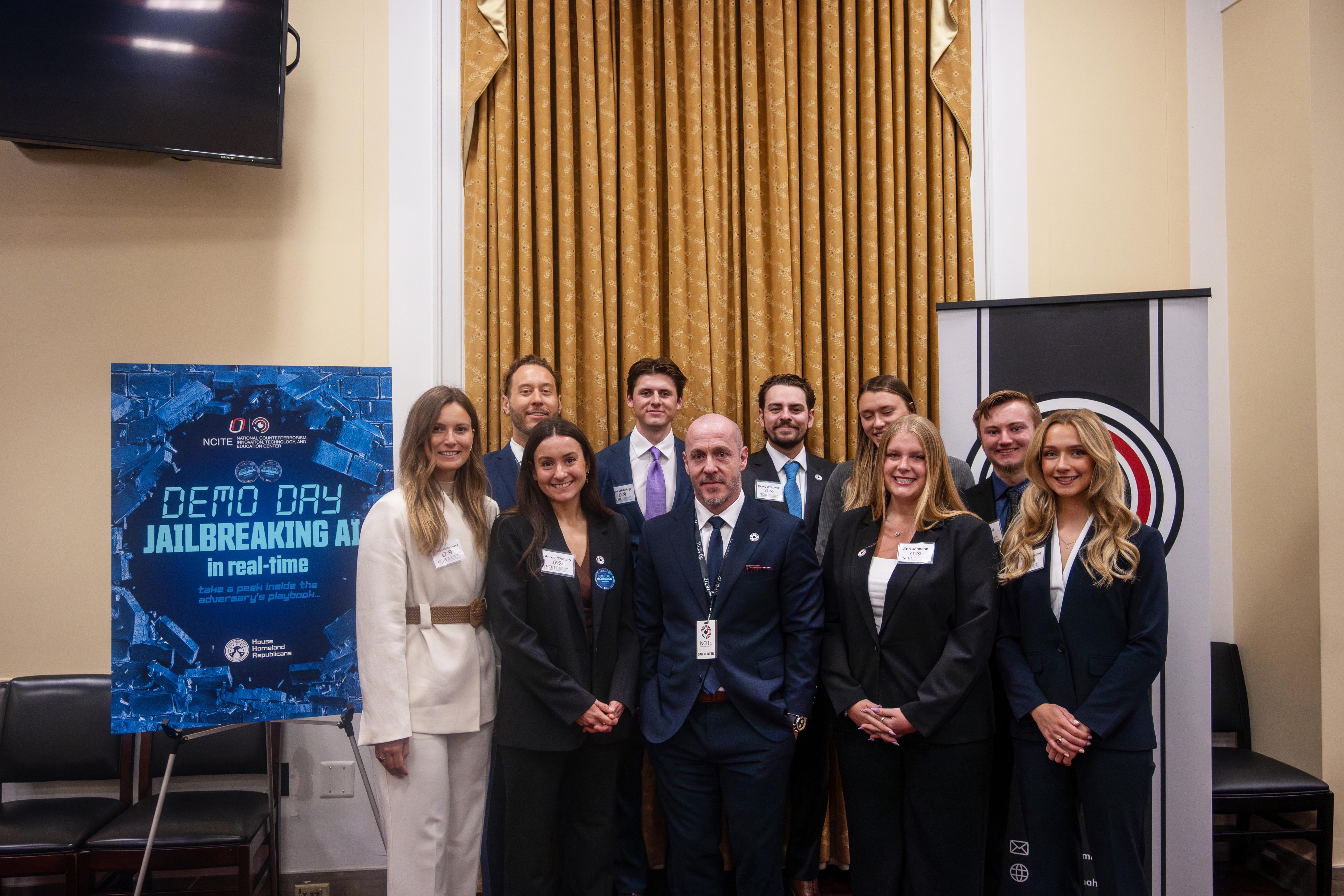A group of professionally dressed participants pose for a photo beside a sign reading “Demo Day: Jailbreaking AI in real-time.”