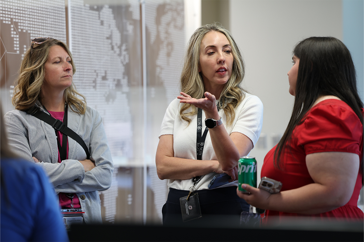 Three women stand together in conversation at an indoor event. The woman in the center gestures with one hand while speaking to the woman on the right, who is holding a can of Sprite.