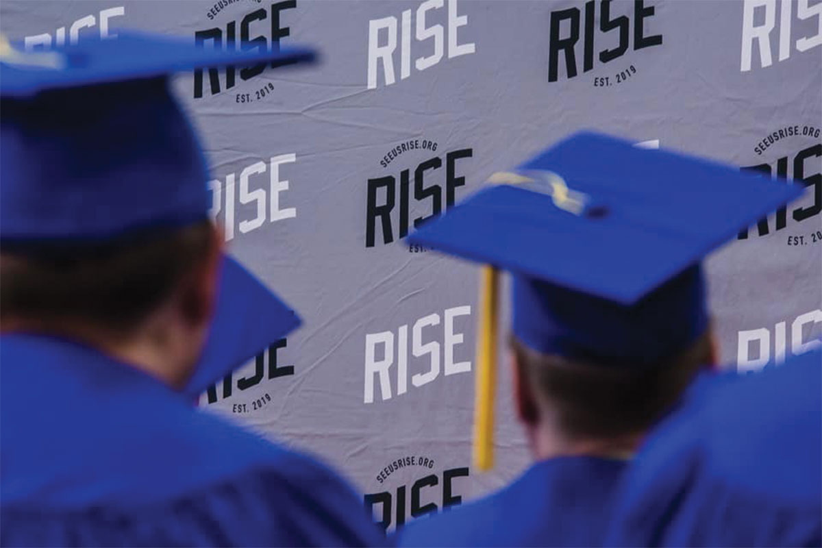 Blurred people wearing blue graduation cap and gowns with RISE Academy backdrop in the foreground.