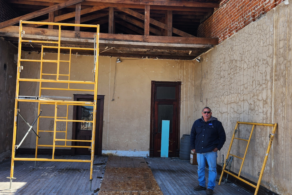 Al standing in one of his renovation projects. The building is old with brick, the roof is missing. Scaffoling is in the room, and it is clearly a worksite. 