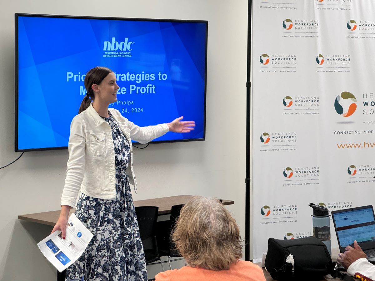 Kiley Phelps presenting a workshop titled 'Pricing Strategies to Maximize Profit' on July 24, 2024, hosted by the Nebraska Business Development Center (NBDC) at Heartland Workforce Solutions. She is standing in front of a large screen and gesturing while holding printed materials, with attendees seated and listening.