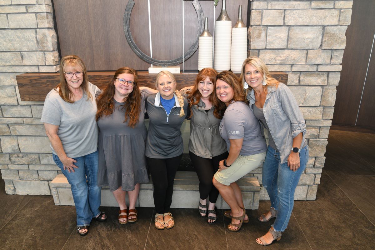 Group photo of six smiling women standing in front of a stone fireplace with decorative vases and a circular sculpture on the mantle. They are dressed casually in gray and blue tones, with some wearing t-shirts branded with 'COB' and 'NBDC.' The group appears friendly and relaxed, posing closely together indoors.
