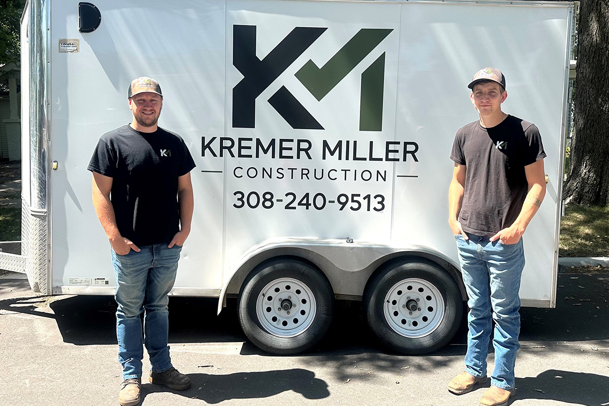 The image shows two men standing in front of a white trailer with the logo and branding for Kremer Miller Construction. The trailer includes their contact phone number, 308-240-9513, and a large logo featuring the letters KM in black and green. Both men are wearing black Kremer Miller Construction t-shirts, jeans, work boots, and caps, and they are smiling at the camera. The setting appears to be outdoors, likely near a work site or residential area with trees providing some shade.