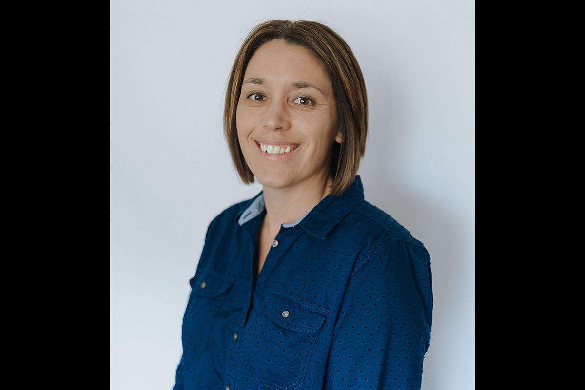 A woman with short brown hair wearing a blue button-up shirt smiles while standing in front of a plain light-colored background.