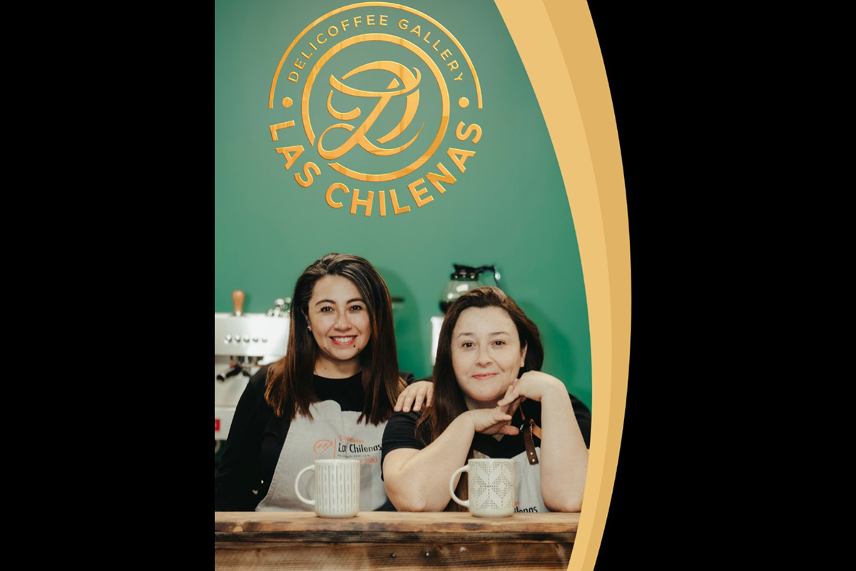 Two women are standing behind a wooden counter in a coffee shop, smiling at the camera. Both are wearing light-colored aprons and holding mugs. Behind them, a green wall displays a large circular gold logo that reads “DELICOFFEE GALLERY LAS CHILENAS” with a stylized letter “L” in the center. Coffee equipment is visible in the background.