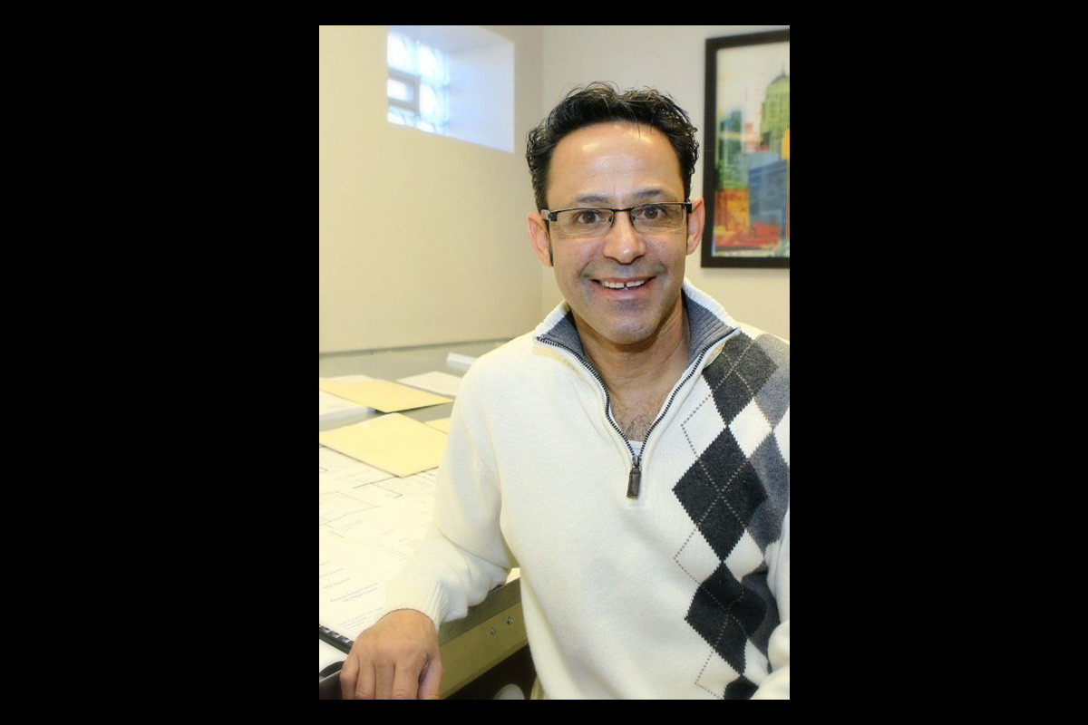 A person wearing glasses and a light-colored sweater with a gray argyle pattern smiles while seated at a desk with papers and folders. A framed colorful cityscape artwork hangs on the wall behind them.