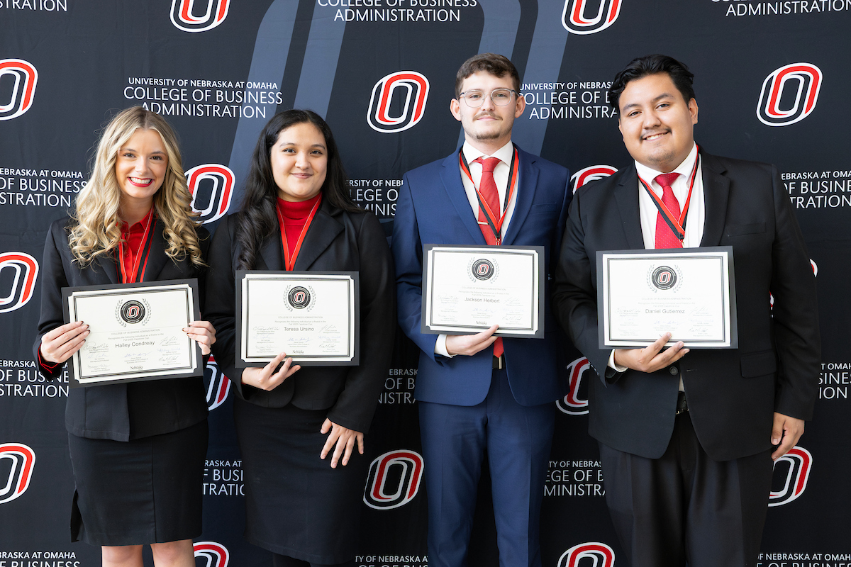 Four students stand in front of a University of Nebraska at Omaha College of Business Administration backdrop, each holding a framed certificate. They are dressed in formal attire with red medals around their necks, smiling for the photo.