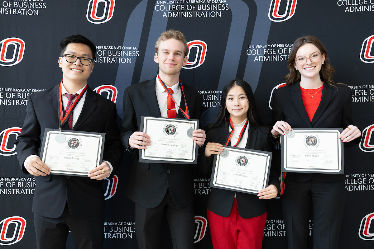 Four students stand in front of a University of Nebraska at Omaha College of Business Administration backdrop, each holding a framed certificate and wearing medals around their necks. They are dressed in formal attire and smiling at the camera.