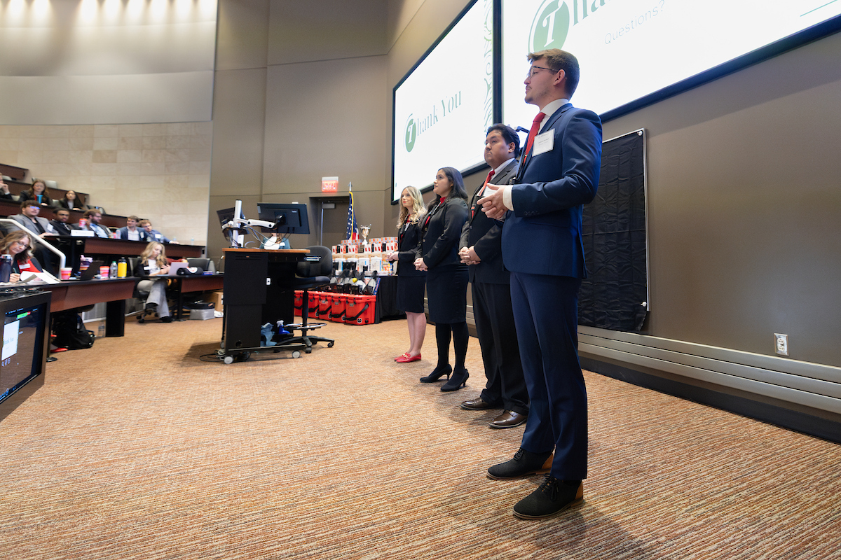 Four students in formal attire stand at the front of a lecture hall giving a presentation. A large screen behind them displays a slide that says “Thank You — Questions?” Audience members sit in tiered seating on the left, and presentation equipment and trophies are visible near the front of the room.