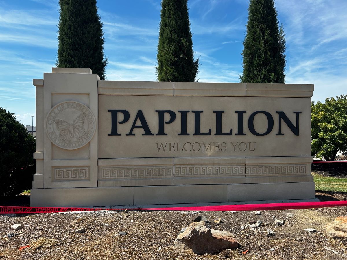 A large cream-colored stone welcome sign reads “PAPILLION WELCOMES YOU.” A circular emblem on the left shows a butterfly and the words “City of Papillion – Founded in 1870.” The sign stands outdoors with tall evergreen trees and a blue sky in the background.