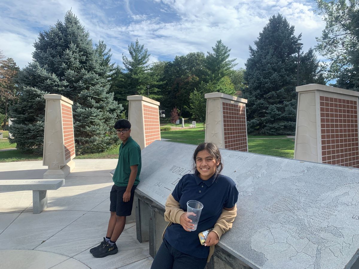 Two kids stand at an outdoor memorial site with tall evergreen trees in the background. One leans against a large engraved stone map while holding a drink cup and smiling. The other stands nearby in front of brick-and-stone pillars. The sky is partly cloudy above the landscaped park area.