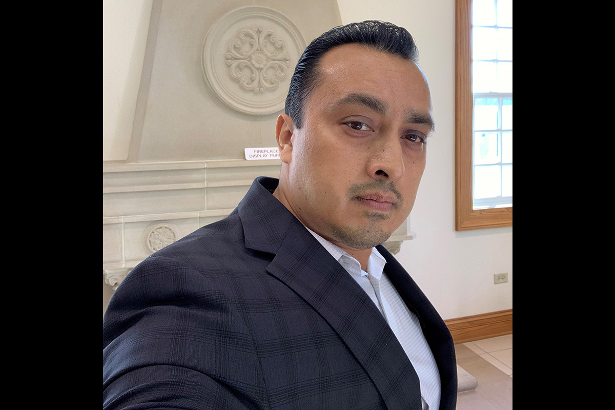 A man in a dark plaid suit jacket takes a selfie indoors. He stands in front of a decorative stone fireplace with an ornate circular carving. A window with wooden trim lets in natural light on the right side of the image.