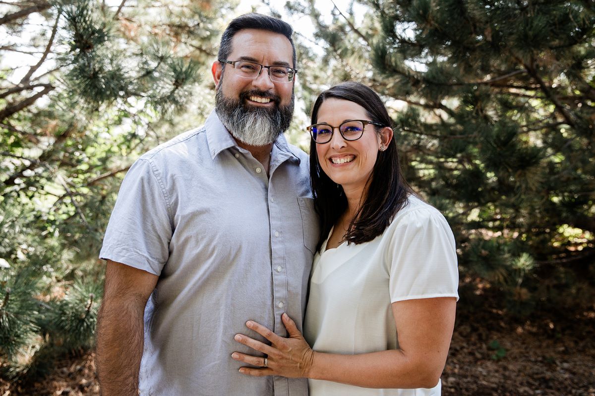 A smiling man and woman stand close together outdoors in front of pine trees. The man has short dark hair, a full beard, and glasses, and is wearing a light gray short-sleeve button-up shirt. The woman has long dark hair, glasses, and is wearing a white blouse with short sleeves. They appear happy and relaxed in a natural setting.