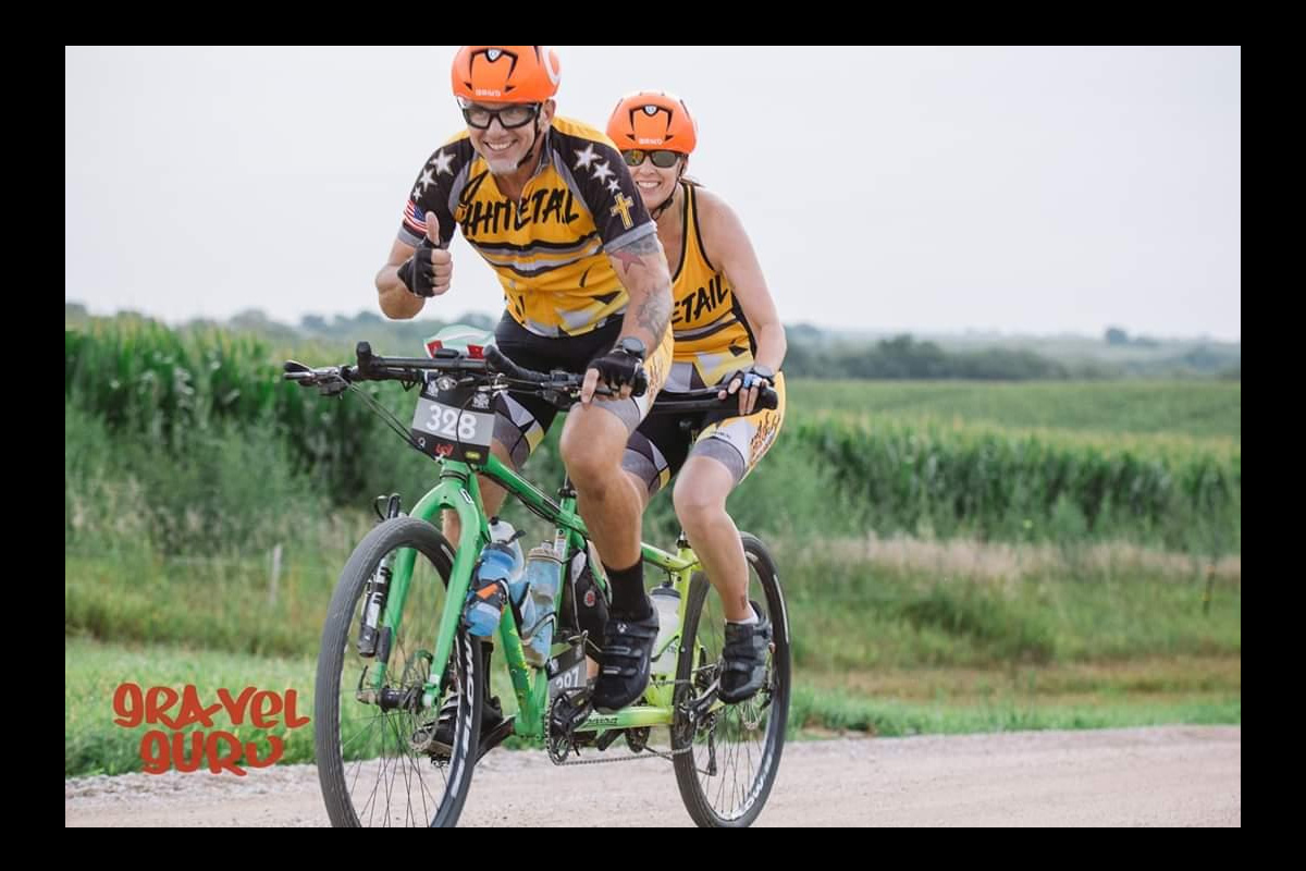 Two cyclists wearing matching yellow jerseys and orange helmets ride a tandem bicycle on a gravel road during a race, smiling and giving a thumbs-up, with green fields in the background.