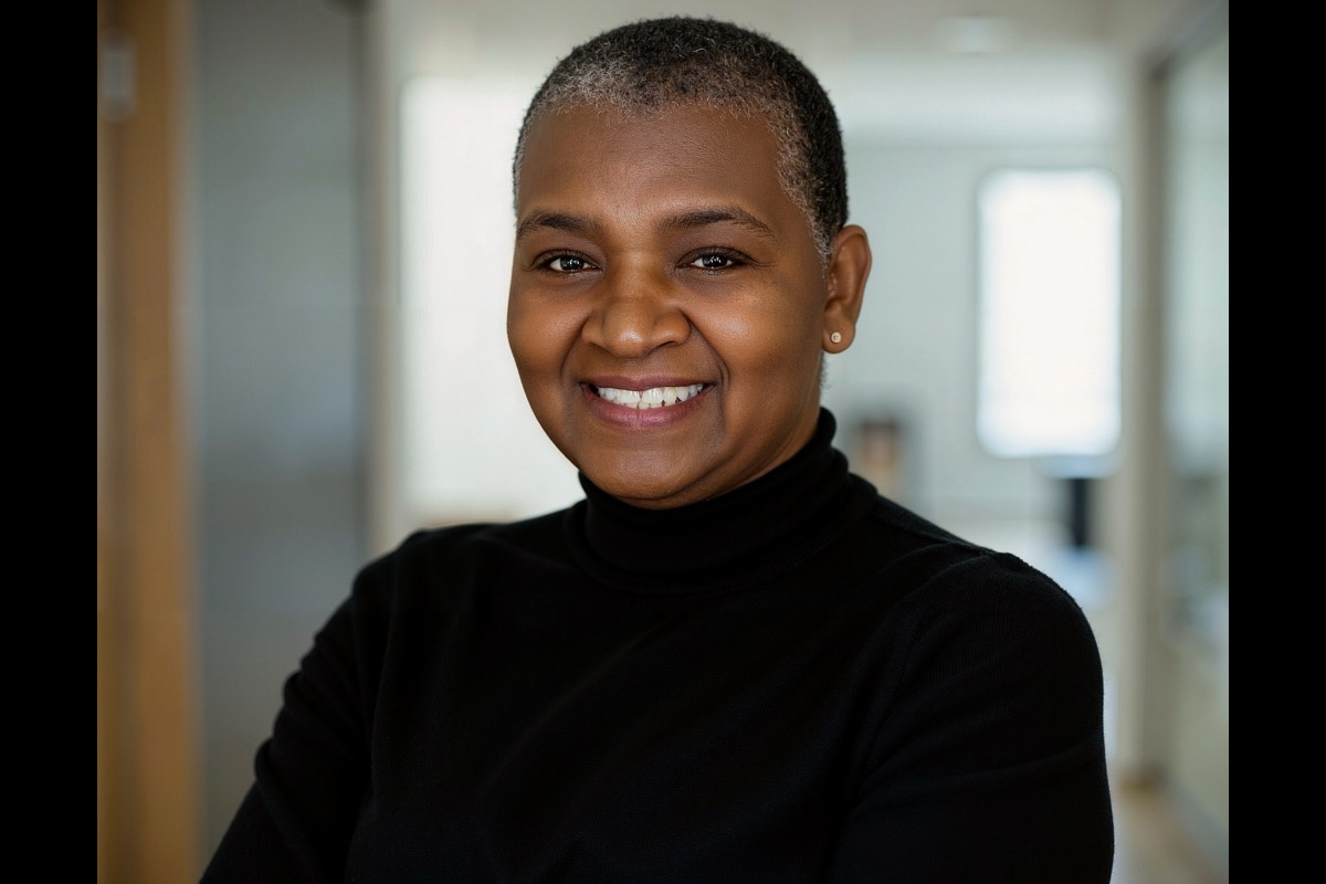 A smiling person with short gray hair wearing a black top, standing indoors in a softly lit hallway with a blurred background. Their arms are crossed, and they are facing the camera.