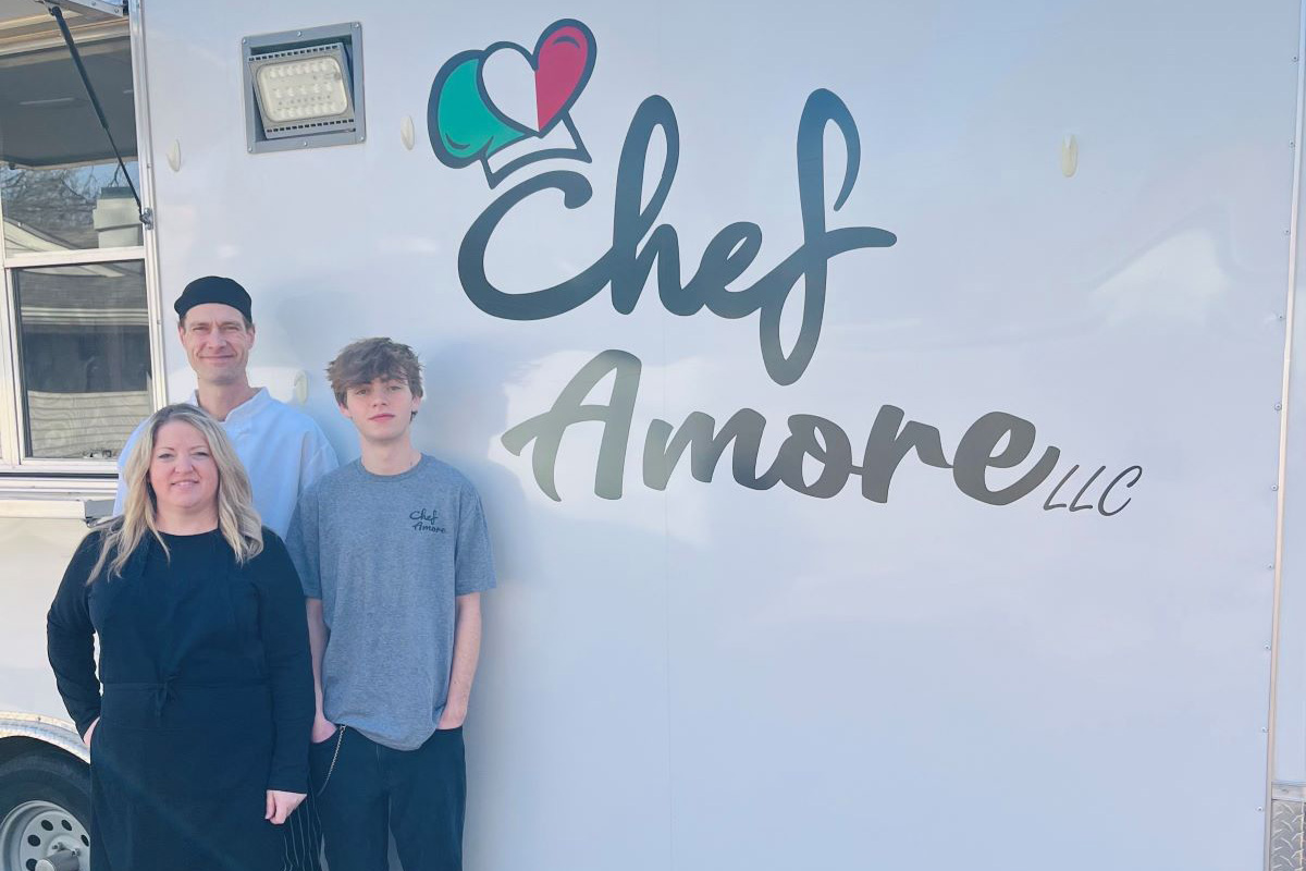 Three people stand outdoors beside a white food truck trailer with the logo “Chef Amore LLC” displayed in large lettering. A woman wearing a black apron stands in front, with a man in a chef’s jacket and hat and a teenage boy in a gray T-shirt standing behind her. The photo appears to be taken on a sunny day in a grassy yard with trees nearby.