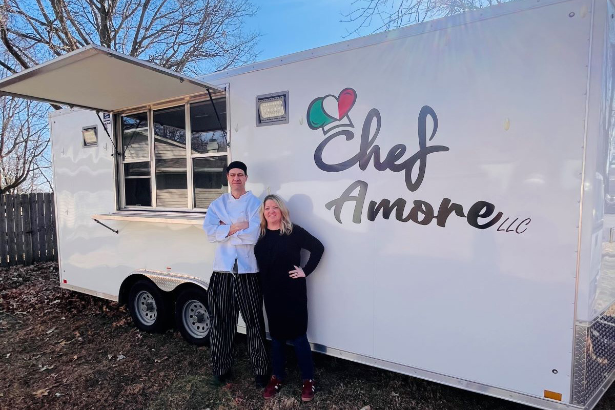 A man and woman stand outdoors beside a white food truck trailer labeled “Chef Amore LLC.” The man wears a white chef’s jacket, black chef’s hat, and striped chef pants, standing with his arms crossed. The woman stands next to him with one hand on her hip, wearing a black top and jeans. The trailer’s service window is open, and the photo is taken on a sunny day in a yard with trees and a wooden fence in the background.
