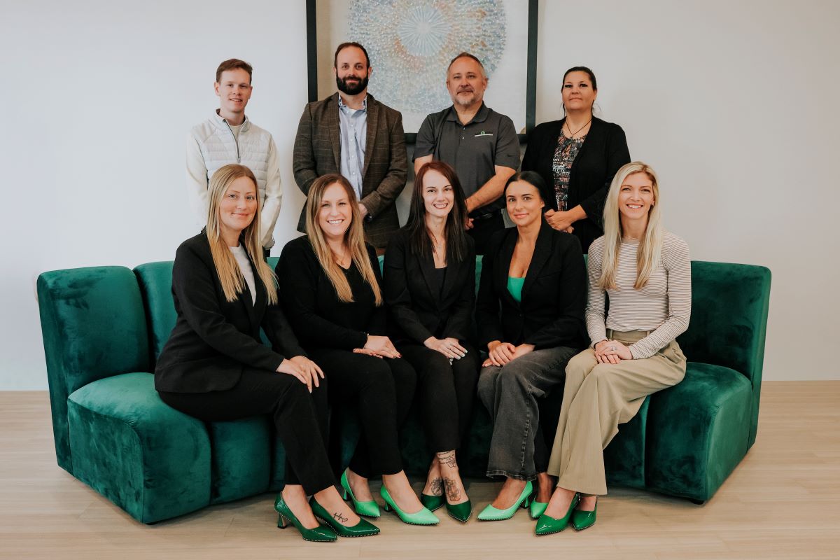 A group of nine professionals pose for a team photo in an office setting, with five seated on a green couch in front and four standing behind them, all dressed in business attire and smiling.