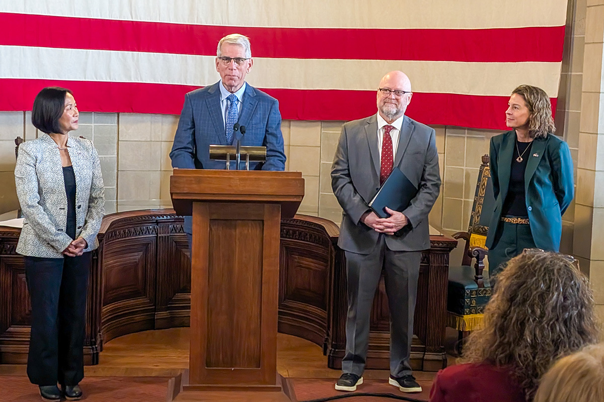 Four officials stand at the front of a formal room beneath a large American flag. A man in a blue suit speaks at a wooden podium with microphones, while three others—two women and one man in business attire—stand beside him listening. Chairs and a wooden desk are visible behind them, and members of the audience can be seen in the foreground.