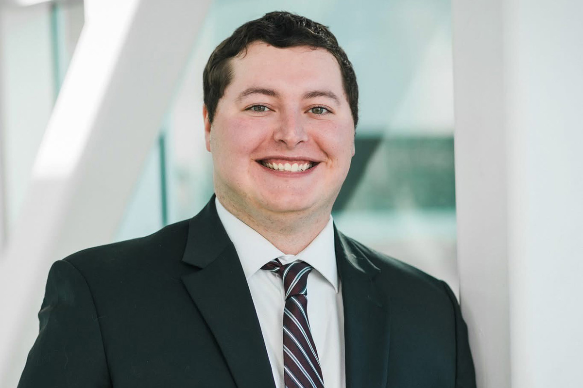 A smiling person wearing a dark suit, white shirt, and striped tie, standing indoors against a softly lit, blurred background.