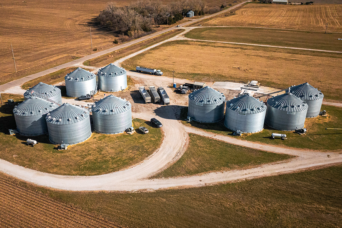 Aerial view of a rural grain storage facility with multiple large metal silos arranged in a cluster, surrounded by dirt roads, farm equipment, and open agricultural fields.
