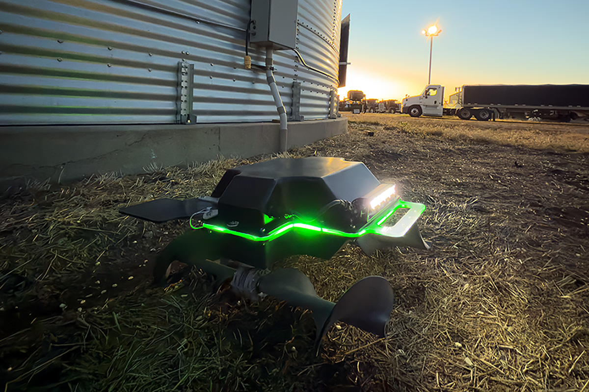 Autonomous agricultural robot with green LED lights sits on the ground beside a metal grain bin at sunset, with semi-trucks and farm equipment visible in the background.