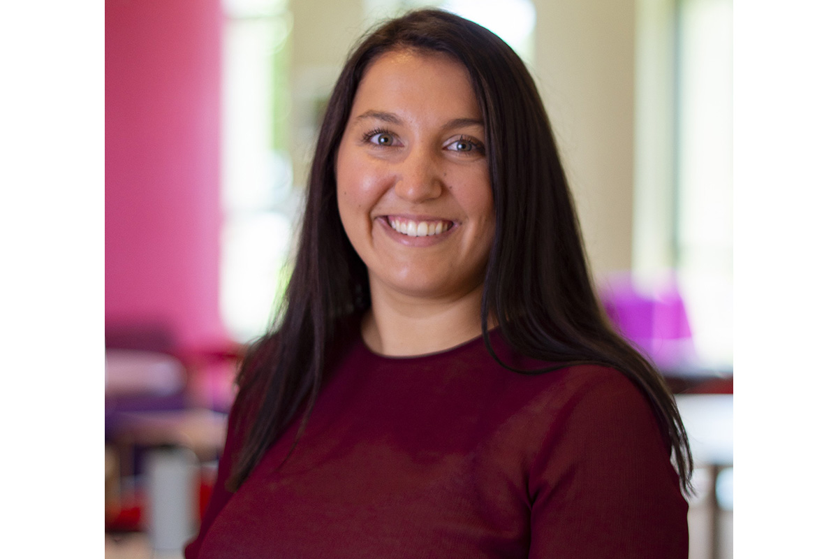 A smiling person with long dark hair wearing a maroon top, standing indoors with a softly blurred, colorful background.