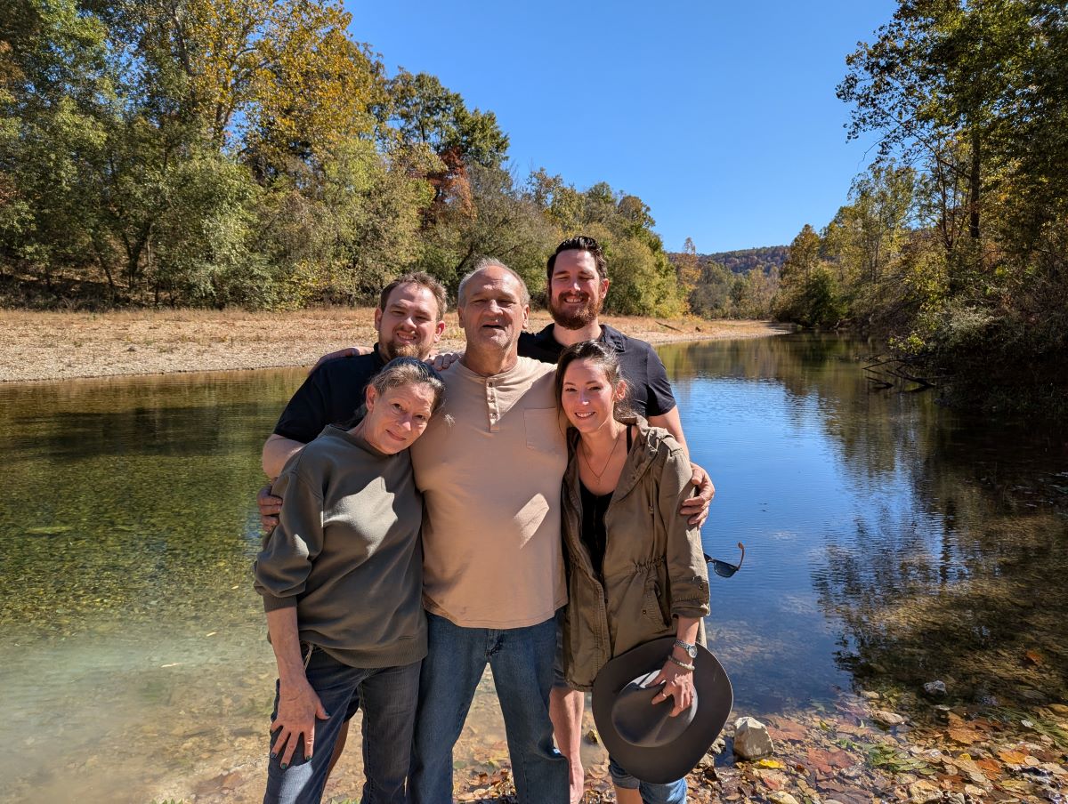 Five family members stand together by a clear river, surrounded by trees with autumn foliage under a bright blue sky.