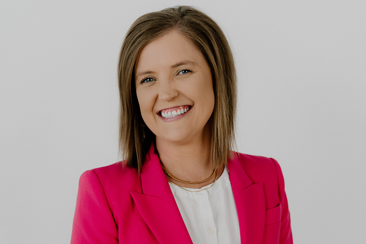 A smiling woman with shoulder-length brown hair wears a bright pink blazer over a white blouse. She is photographed against a light gray background in a professional headshot.