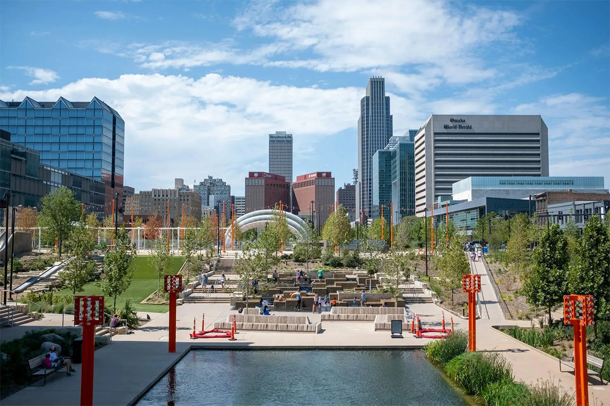 A wide daytime view of downtown Omaha, Nebraska, seen from a landscaped urban park. In the foreground, a reflective pool borders paved walkways, terraced seating, trees, and bright orange light poles. Small groups of people are scattered throughout the park. The city skyline rises in the background, including the WoodmenLife Tower, the First National Bank Tower, and the Omaha World-Herald building, beneath a blue sky with scattered white clouds.