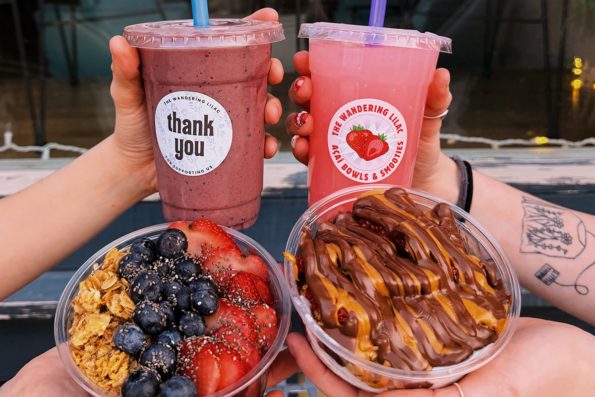 Two hands hold colorful smoothies from The Wandering Lilac in front of a storefront window displaying business hours.