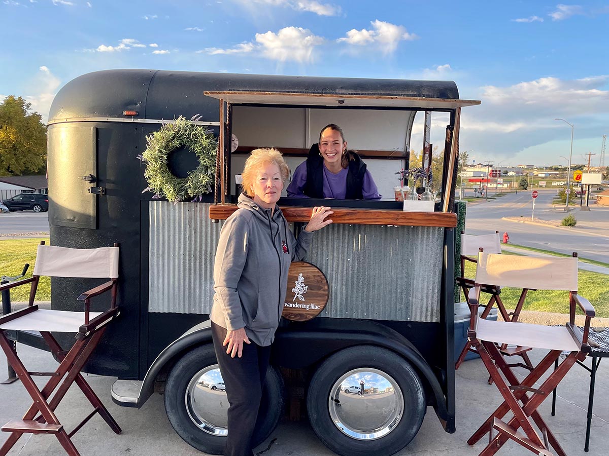 Two women pose with The Wandering Lilac mobile food trailer, with one standing outside and the other smiling through the service window.