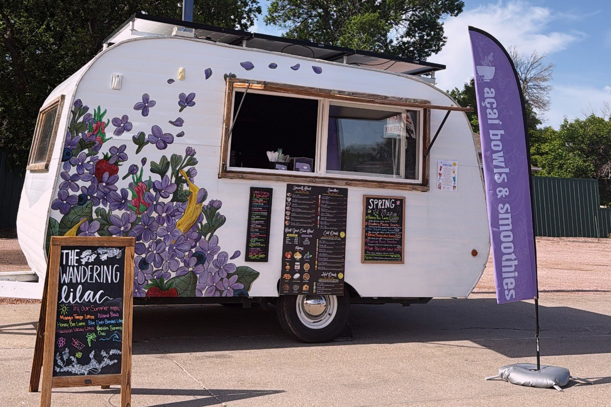 A white food trailer decorated with colorful lilac flowers displays “The Wandering Lilac” signage and menus for açaí bowls and smoothies.