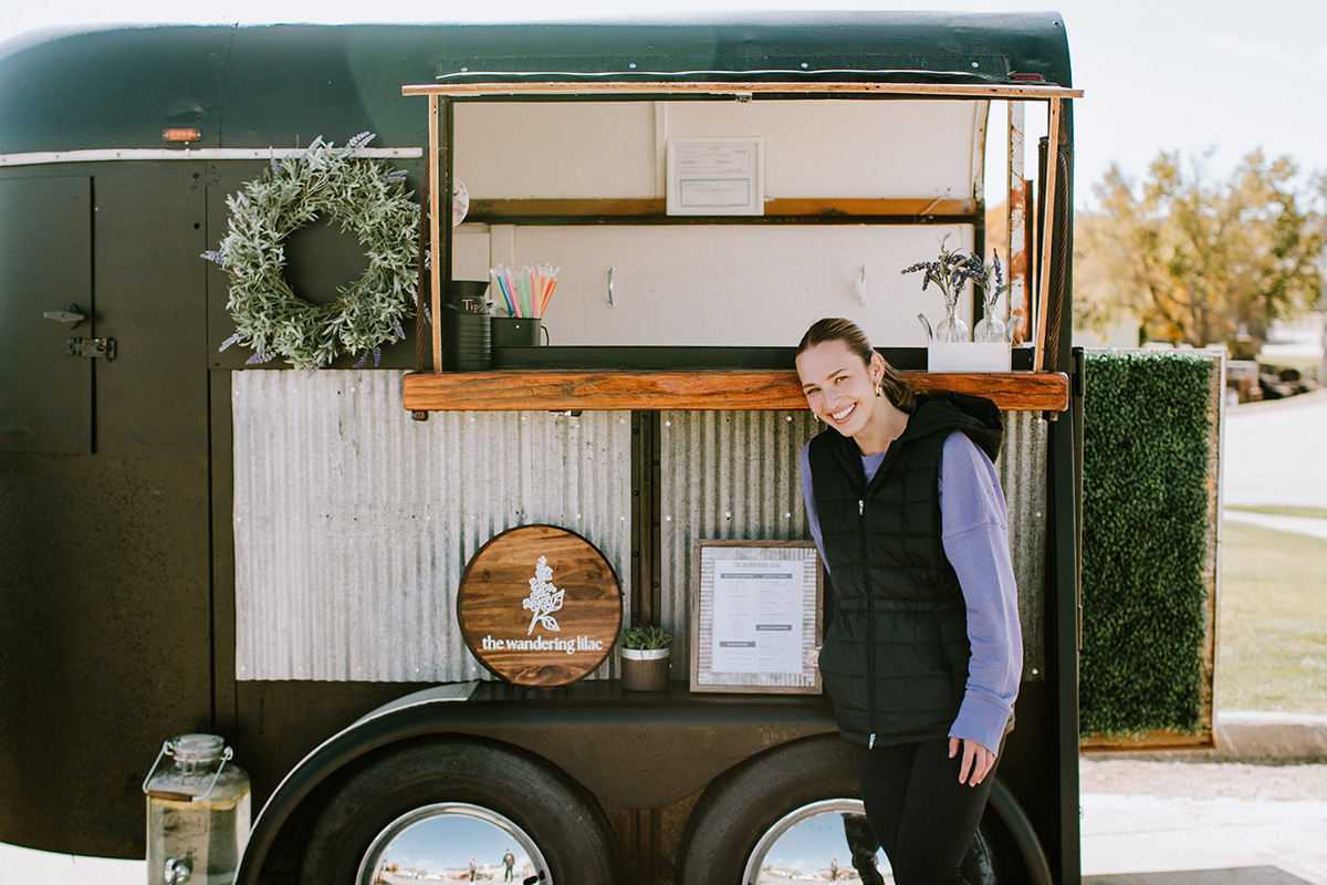 A smiling woman stands beside The Wandering Lilac mobile food trailer, posing in front of its open service window and business sign.